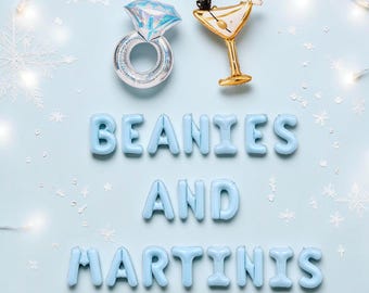 May include: Light blue foil balloons spelling the text BEANIES AND MARTINIS arranged in three rows. Above the text are two foil balloons: one shaped like a diamond ring with a silver band and one shaped like a martini glass with gold accents.