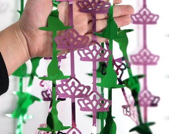 May include: Green witch hat and purple crown-shaped hanging decorations. The decorations are made of shiny, reflective material. The witch hats are green and the crowns are purple. The decorations are held by a hand.