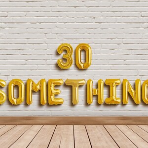 May include: Gold foil balloon letters and numbers spelling "30 SOMETHING" against a white brick wall and wooden floor. The balloons are arranged to celebrate a milestone birthday.