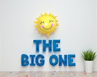 May include: A yellow sun-shaped balloon with a smiling face and the text "THE BIG ONE" in blue foil balloons. A potted plant sits to the right. Colorful confetti dots the floor.