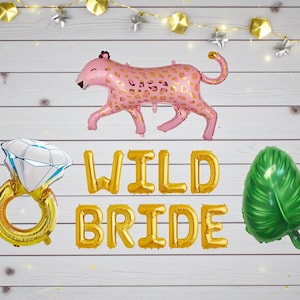 May include: A set of foil balloons for a bridal celebration. Features a pink leopard, a diamond ring, a green leaf, and gold letter balloons spelling "WILD BRIDE". Fairy lights are strung across the top.