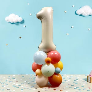 May include: A festive birthday decoration featuring a large beige number one balloon atop a cluster of colorful balloons. The arrangement includes red, orange, yellow, and blue balloons. The background is light blue with cloud and star decorations.