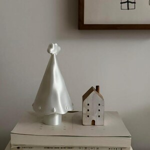 Puede incluir: Un objeto cónico blanco y liso en forma de árbol con una pequeña estrella en la punta y una pequeña figura de madera blanca en forma de casa con una chimenea marrón y ventanas cuadradas, ambos sobre una pila de libros blancos.