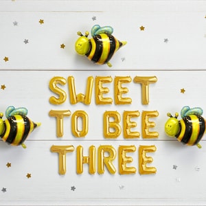 May include: A festive birthday arrangement with three bee-shaped balloons and gold foil letter balloons spelling "SWEET TO BEE THREE". The background is white with scattered gold and silver star confetti.