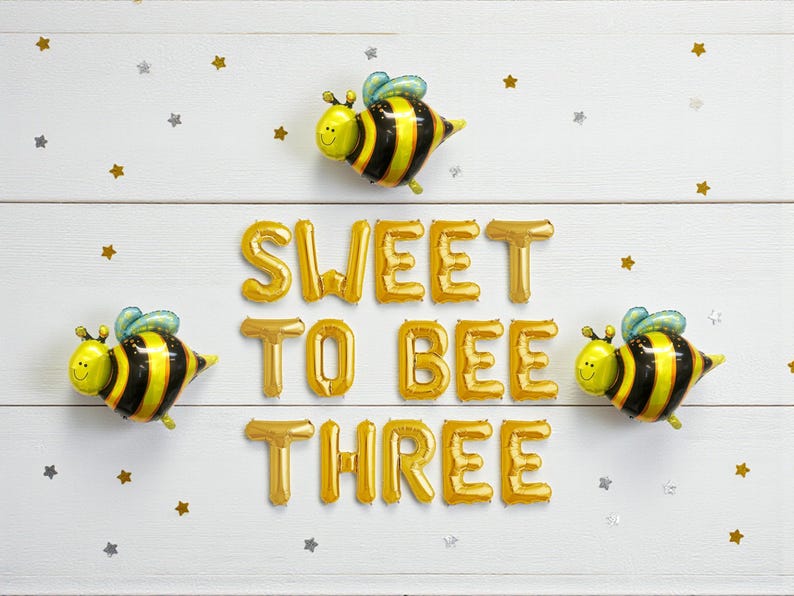 May include: A festive birthday arrangement with three bee-shaped balloons and gold foil letter balloons spelling "SWEET TO BEE THREE". The background is white with scattered gold and silver star confetti.