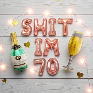 May include: A celebratory arrangement with rose gold foil balloons spelling "SHIT IM 70", a champagne bottle balloon, and a champagne glass balloon. Gold heart confetti and string lights are scattered on a white wood background.