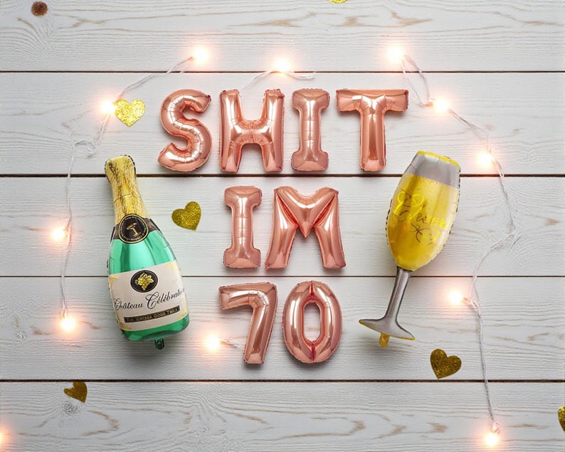 May include: A celebratory arrangement with rose gold foil balloons spelling "SHIT IM 70", a champagne bottle balloon, and a champagne glass balloon. Gold heart confetti and string lights are scattered on a white wood background.