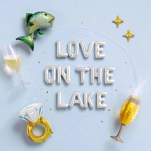 May include: A celebratory scene with foil balloons spelling "LOVE ON THE LAKE." The image includes a fish balloon, champagne glasses, a diamond ring balloon, and star-shaped balloons, set against a light blue backdrop.