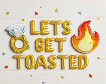 May include: Gold foil balloons spelling "LET'S GET TOASTED" with a diamond ring balloon and a flame balloon. Confetti in various colors is scattered around the balloons on a white wooden surface. Party decorations.