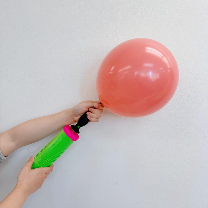 Op de afbeelding: Een koraalkleurige ballon wordt opgeblazen met een groene en roze handpomp. De ballon wordt op zijn plaats gehouden terwijl de pomp wordt gebruikt. De achtergrond is een effen witte muur.