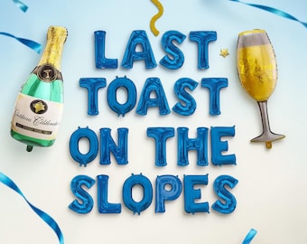 May include: A festive arrangement featuring blue foil balloon letters spelling "LAST TOAST ON THE SLOPES". A champagne bottle and a champagne flute balloon are also present, along with blue ribbons and gold confetti.