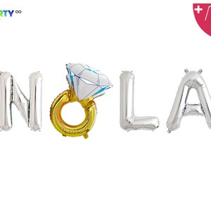 May include: Silver foil balloons spelling out "NOLA" with a gold ring balloon featuring a diamond-shaped top. The balloons are for a party or celebration. The image also includes a balloon straw and string.