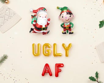 May include: Foil balloons shaped like Santa Claus and an elf, accompanied by gold letter balloons spelling UGLY and red letter balloons spelling AF. The balloons are arranged on a light background with scattered pine needles and confetti.