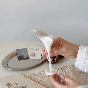 May include: A white, slender, trumpet-shaped vase with a smooth, matte finish. The vase features a flared, organic opening at the top that tapers down into a thin, delicate stem, ending in a small, circular base for stability on flat surfaces.