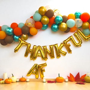 Peut inclure: Décoration festive de Thanksgiving avec une arche de ballons orange, turquoise, dorés et marron. Des ballons dorés forment l'inscription "THANKFUL AF". Citrouilles, bougies et feuilles d'automne complètent la scène.