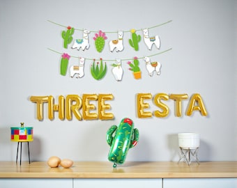 May include: A festive party scene with gold foil balloons spelling "THREE ESTA". Decorative hanging llama and cactus cutouts adorn the wall. A green cactus balloon and other decorative items sit on a wooden surface.