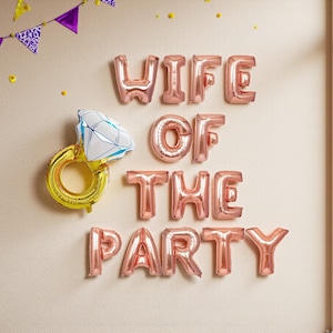 以下が含まれることがあります： 「WIFE OF THE PARTY」と書かれたローズゴールドの風船が壁に飾られ、ダイヤモンド型の宝石が付いた金の指輪の風船も添えられています。紫と白のペナントバナーと黄色の紙吹雪が、お祝いの装飾を完成させています。