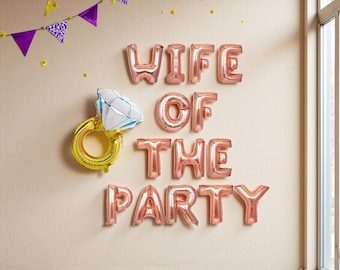 May include: Rose gold foil balloons spelling "WIFE OF THE PARTY" are displayed on a wall, alongside a gold ring balloon with a diamond-shaped gem. Purple and white pennant banner and yellow confetti complete the festive decor.