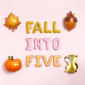 Puede incluir: Globos de aluminio que forman la frase FALL INTO FIVE en dorado, rosa y melocotón. Incluye una hoja de arce dorada, una bellota marrón, una calabaza naranja y un globo de ardilla beige. Todo sobre un fondo de color rosa claro.