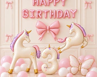 May include: A set of pink and cream foil balloons featuring the text HAPPY BIRTHDAY, a large bow, two unicorns, a number 3, and a butterfly. The display includes several round pink and white balloons arranged on the floor against a wall.