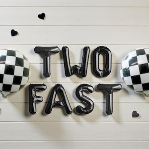 May include: Black foil balloons spelling "TWO FAST" with two checkered racing ballons and black heart decorations on a white wooden background. A festive theme for a birthday or celebration.