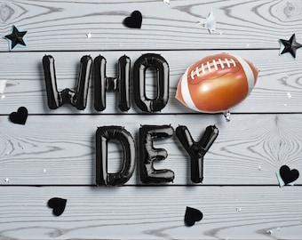 May include: Black foil balloons spelling "WHO DEY" with a football-shaped balloon and scattered black hearts and stars on a light wood background. A festive arrangement for a sports-themed celebration.