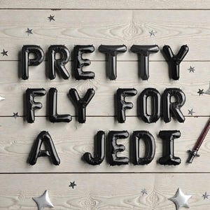 May include: Black balloon letters spell out "PRETTY FLY FOR A JEDI" on a light wood background, with star-shaped and star-burst silver decorations. A lightsaber prop is included.