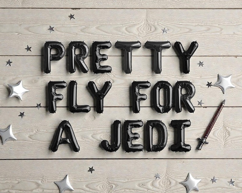 May include: Black balloon letters spell out "PRETTY FLY FOR A JEDI" on a light wood background, with star-shaped and star-burst silver decorations. A lightsaber prop is included.