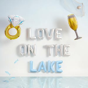 May include: A festive arrangement of foil balloons spelling "LOVE ON THE LAKE." The display includes a gold ring balloon with a diamond, silver letter balloons, and a champagne glass balloon. Light blue fish decorations add to the lake theme.