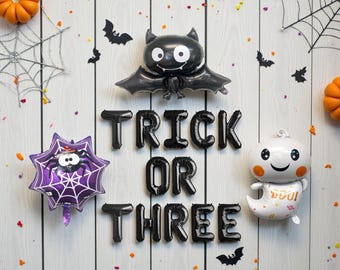 以下が含まれることがあります： 「TRICK OR THREE」と書かれた黒い風船、コウモリの形をした風船、クモの巣の風船、お化けの風船が特徴のハロウィーンをテーマにしたアレンジメント。オレンジ色のカボチャとコウモリの切り抜きが白い木の背景に散らばっています。