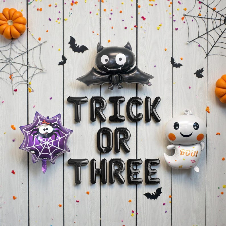 May include: A Halloween-themed arrangement featuring black balloons spelling "TRICK OR THREE", a bat balloon, a spider web balloon, and a ghost balloon. Orange pumpkins and bat cutouts are scattered on a white wooden background.