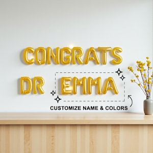May include: Gold foil balloon letters spelling "CONGRATS DR EMMA" on a white wall. The text "CUSTOMIZE NAME & COLORS" is below the name. A vase of yellow flowers sits on a wooden table.