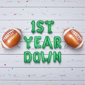 May include: A festive arrangement featuring two football-shaped balloons and green letter balloons spelling "1ST YEAR DOWN." The football balloons are brown with white laces. The background is a white wooden surface with colorful confetti.
