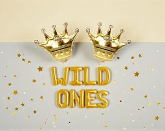 Könnte beinhalten: Zwei goldene Kronenballons und die Worte "WILD ONES" in goldenen Ballonbuchstaben werden auf einem grau-beigen Hintergrund mit goldenen Sternenkonfetti angezeigt.