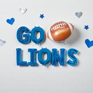 May include: A festive arrangement with blue foil balloons spelling "GO LIONS" and a football-shaped balloon. Silver and blue heart and star decorations are scattered on a white wooden background. Perfect for a sports-themed party.