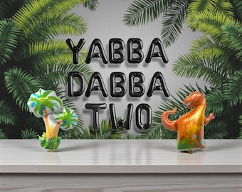 Puede incluir: Globos de papel de aluminio negros deletrean "YABBA DABBA TWO" sobre una superficie blanca. Una palmera y un globo con forma de dinosaurio flanquean el texto. El fondo presenta un exuberante follaje verde, creando una escena festiva.