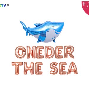 May include: A blue and white shark-shaped balloon above rose gold foil letter balloons spelling "ONE DER THE SEA". The balloons are for a party. Includes a balloon straw and string.