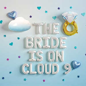 May include: A light blue wall decorated with silver foil balloons spelling "THE BRIDE IS ON CLOUD 9". The scene includes a cloud-shaped decoration, a diamond ring balloon, and heart and star-shaped confetti.