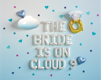 May include: A light blue wall decorated with silver foil balloons spelling "THE BRIDE IS ON CLOUD 9". The scene includes a cloud-shaped decoration, a diamond ring balloon, and heart and star-shaped confetti.