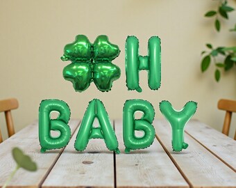 May include: Green foil balloons spelling "BABY" and a four-leaf clover, plus the letter "H". The balloons are arranged on a wooden table. The background includes a light-colored wall and greenery.