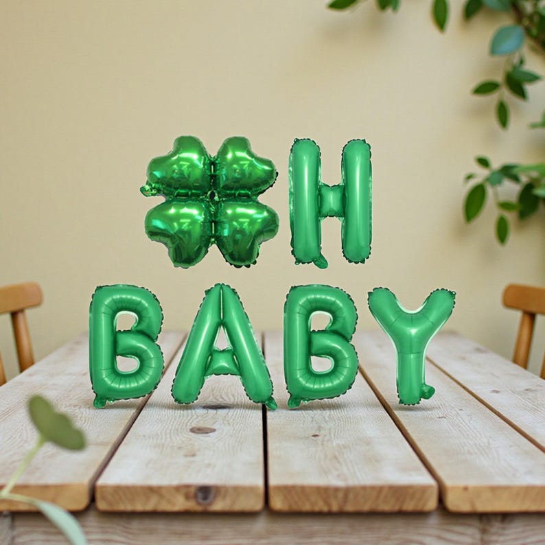 May include: Green foil balloons spelling "BABY" and a four-leaf clover, plus the letter "H". The balloons are arranged on a wooden table. The background includes a light-colored wall and greenery.