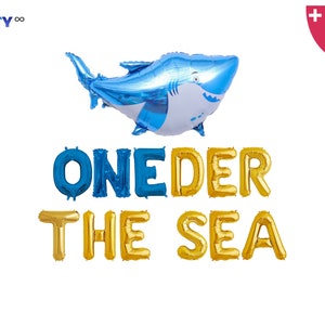 May include: A blue and white shark-shaped balloon above gold and blue letter balloons spelling "ONEDER THE SEA". Includes a balloon straw and string. Party decor for a first birthday celebration.