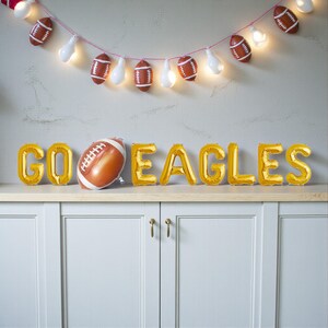 以下が含まれることがあります： 「GO EAGLES」と書かれた金色の風船文字と、フットボール型の風船を使ったお祝いの飾り付け。電飾とフットボール型のオーナメントが上に吊り下げられています。ニュートラルな背景に配置されており、ゲームデーのお祝いに最適です。
