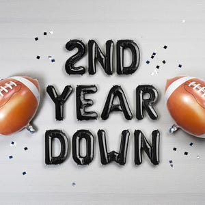 Puede incluir: Vista aérea de dos globos con forma de balón de fútbol americano y globos con letras negras que deletrean "2ND YEAR DOWN". Los balones de fútbol son marrones con rayas y cordones blancos. Fondo gris claro con confeti.