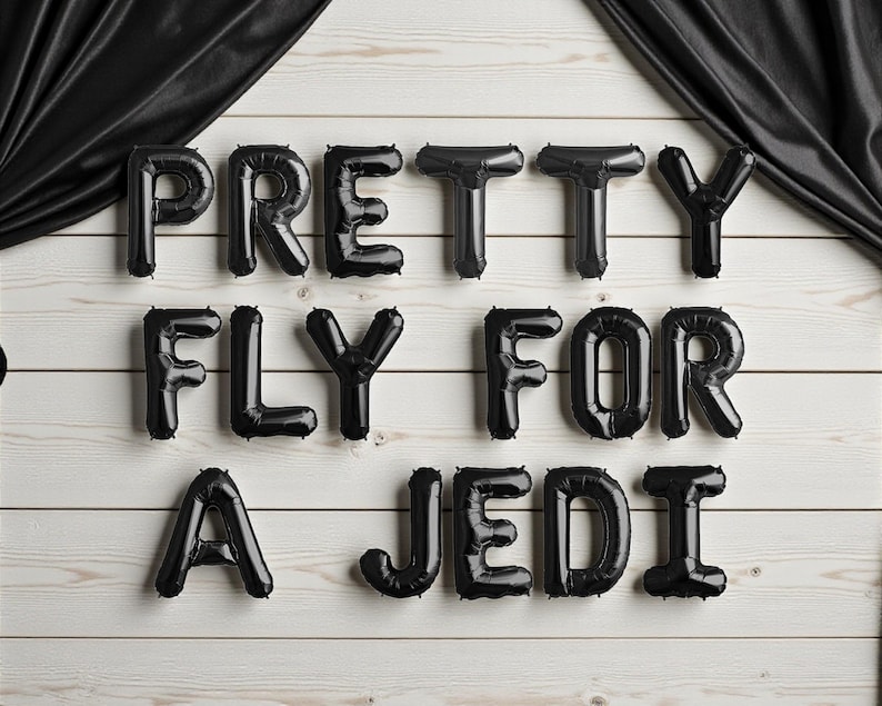 May include: Black balloon letters spelling out "PRETTY FLY FOR A JEDI" against a white wooden background. The balloons are arranged in three rows, with the phrase's letters in a bold, sans-serif font. Black fabric drapes from the top corners.