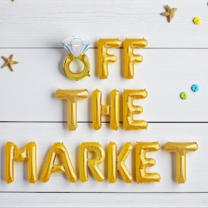 May include: Gold foil balloon letters spell "OFF THE MARKET" with a diamond ring balloon. The balloons are arranged on a white wooden background, decorated with starfish and floral accents. A festive engagement or wedding theme.