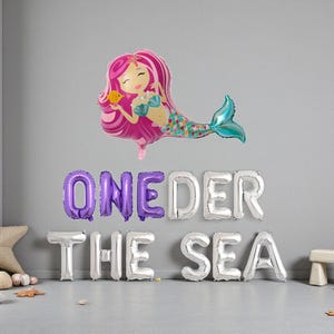 Op de afbeelding: Een ballon in de vorm van een zeemeermin met roze haar en een turquoise staart zweeft boven zilveren en paarse letterballonnen met de tekst "ONEDER THE SEA". Een zeester en een kleine houten kruk staan op de grond.