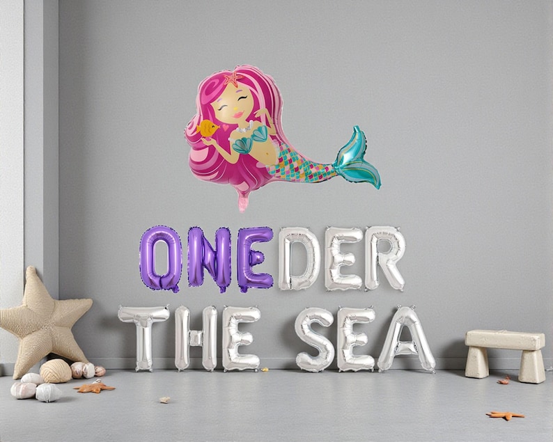 Op de afbeelding: Een ballon in de vorm van een zeemeermin met roze haar en een turquoise staart zweeft boven zilveren en paarse letterballonnen met de tekst "ONEDER THE SEA". Een zeester en een kleine houten kruk staan op de grond.
