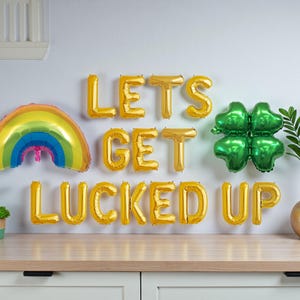 Puede incluir: Una festiva exhibición del Día de San Patricio con globos de letras doradas que deletrean "LET'S GET LUCKED UP". También se incluye un globo de arcoíris y un globo de trébol verde. Un jarrón dorado con vegetación y pequeñas plantas en macetas completan la escena.