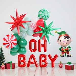 May include: A festive arrangement of red, green, and white balloons, including star-shaped and candy cane designs. The phrase "OH BABY" is spelled out in red balloons, alongside an elf-shaped balloon. Small wrapped gifts and a Christmas tree are also present.